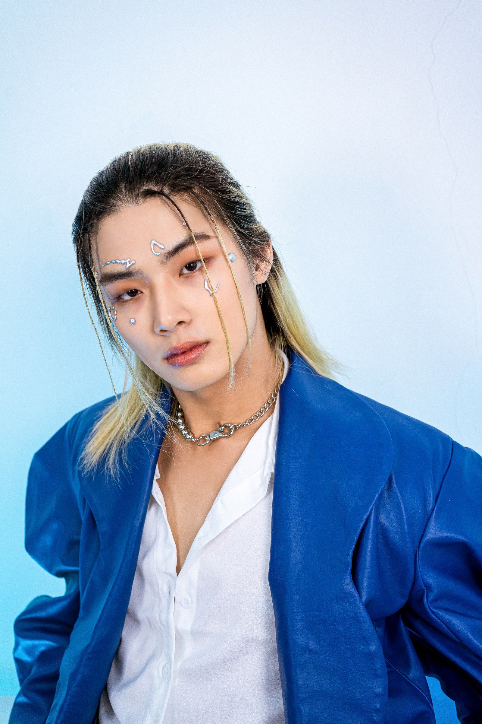 白いシャツに光沢のある青いジャケットを羽織ったTaichan。顔にはシルバーの装飾、首元にはチェーンネックレスを着け、淡いブルーの背景の前でカメラを見つめています。